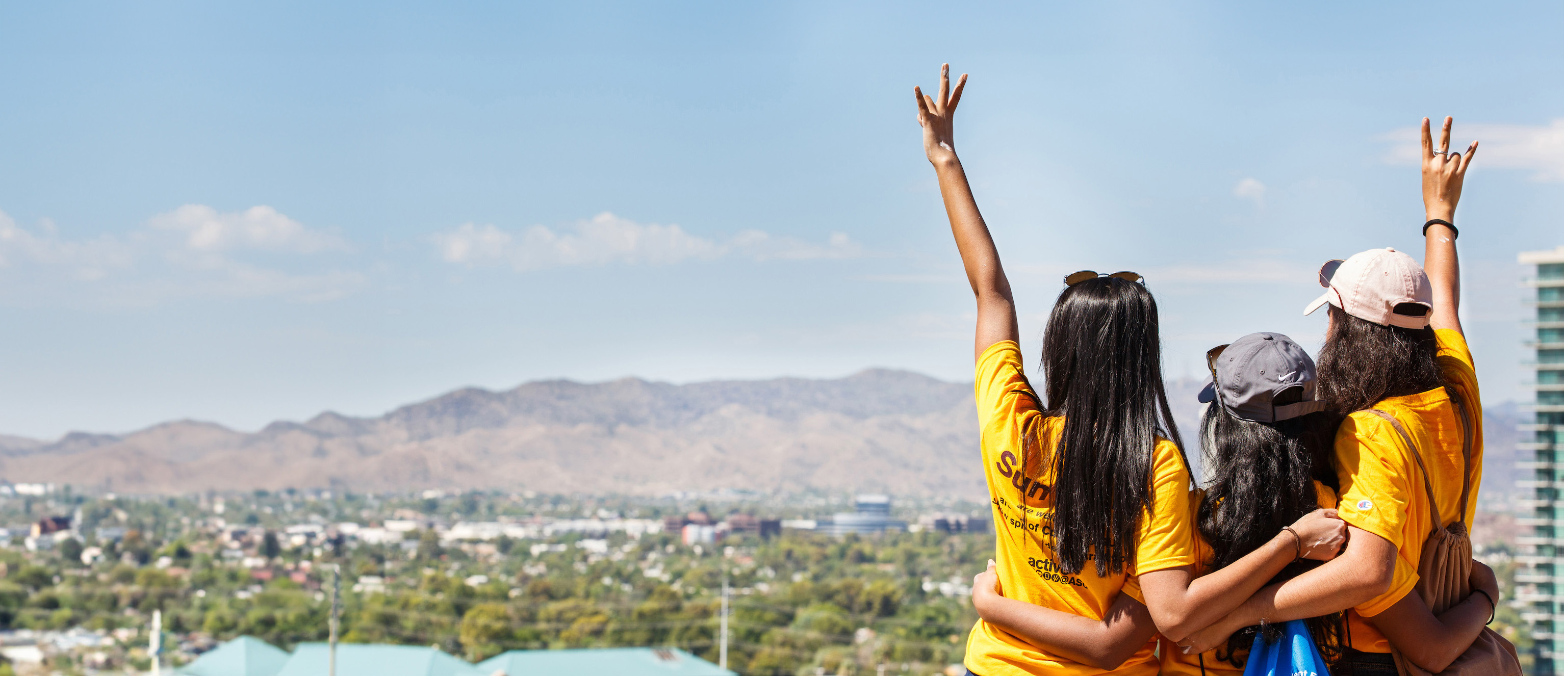 A-S-U students with their backs turned, looking over a cityscape and holding up the pitchfork sign with their hands